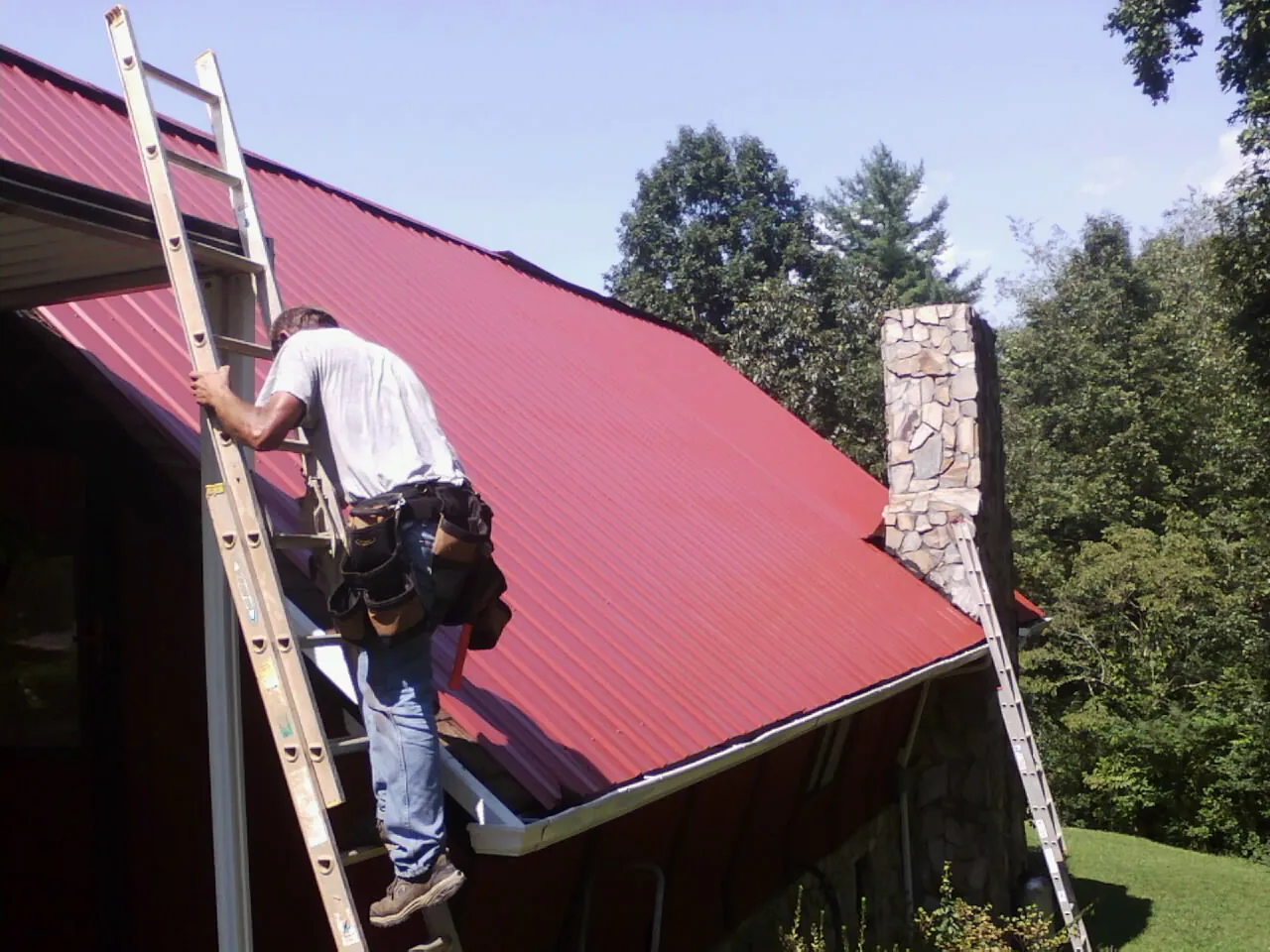 Roofer on a ladder beside a red metal roof and stone chimney — Soffit & Fascia Repair by Roofing Brothers in Estacada
